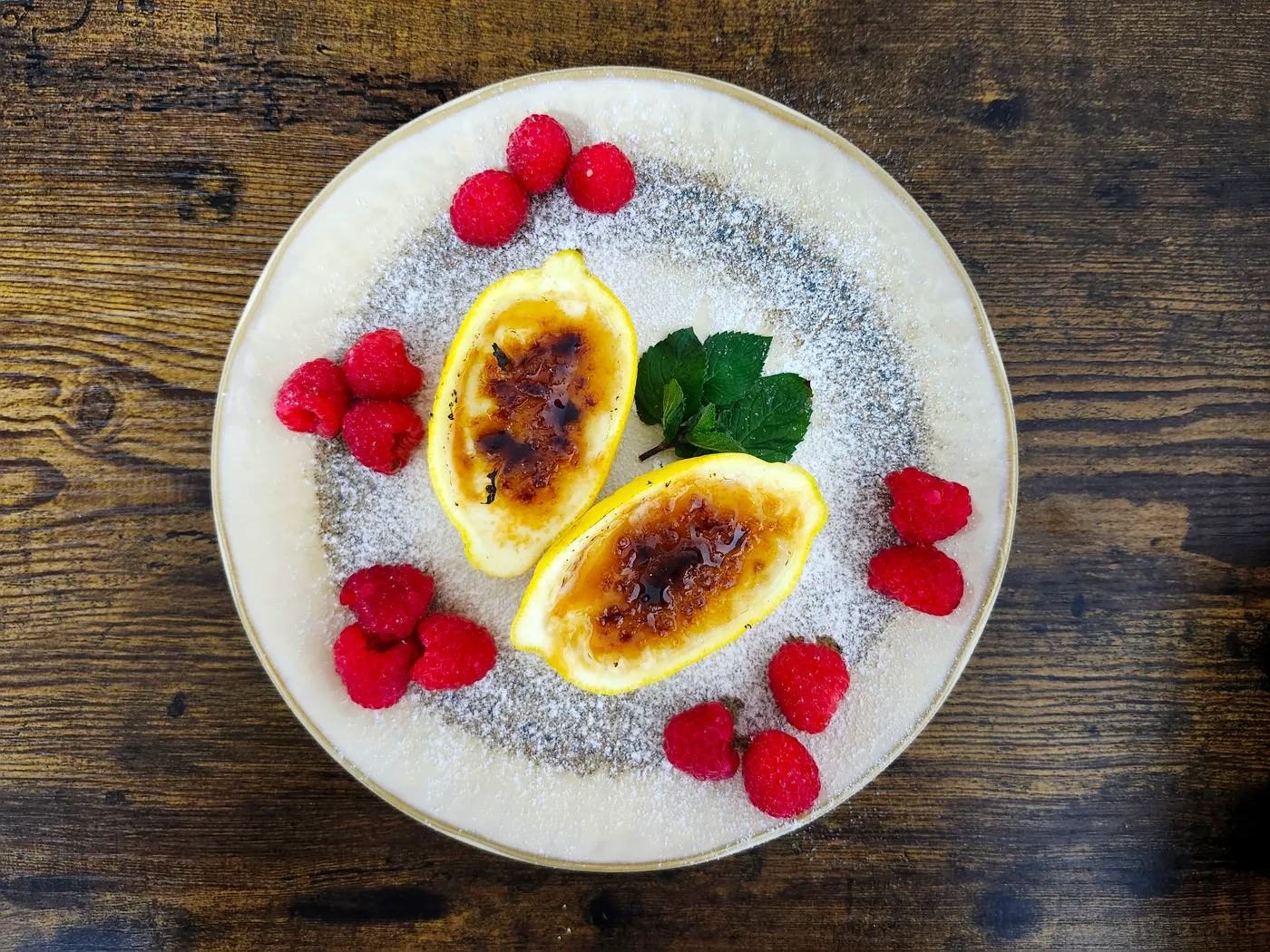 Zwei Lemon Posset Brûlée in ausgehöhlten Zitronenhälften auf einem Teller mit Himbeeren, Minze und Puderzucker